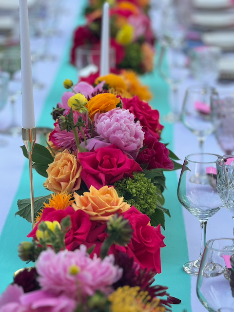 Stunning table centerpiece featuring rich mix of roses, peonies, ranunculus in vibrant pinks, oranges, and yellows with elegant table styling